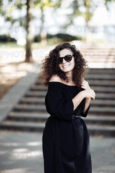 Beautiful young curly woman in a black dress and sunglasses posing and smiling in the city