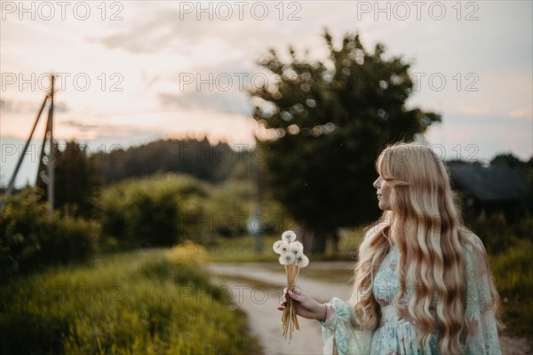 A beautiful young blonde woman with long hair in a white dress walks through a field of dandelions