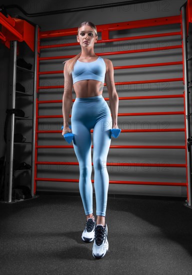 Beautiful tall blonde posing in the gym with dumbbells in her hands against the background of the wall bar. The concept of sports