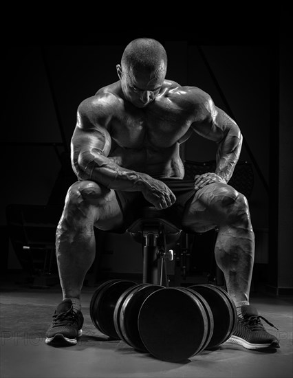 Muscular man sits on a bench near huge dumbbells. Bodybuilding and powerlifting concept.