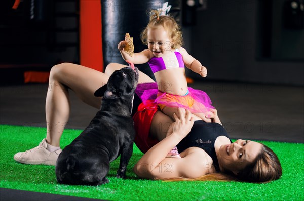 Charming sports mom plays in the gym with her little daughter and a French bulldog.