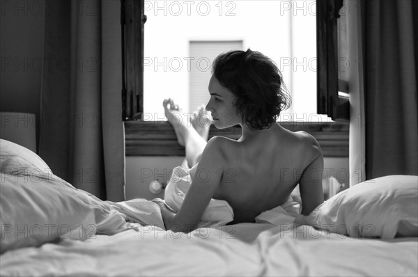 Charming girl lies in bed in the morning. She stretched her legs to the window and looks at the camera over her shoulder. The concept of relaxation