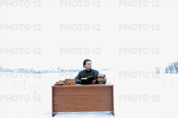 A young handsome intelligent man in a jacket sits at a table in a field and reads ancient books in winter