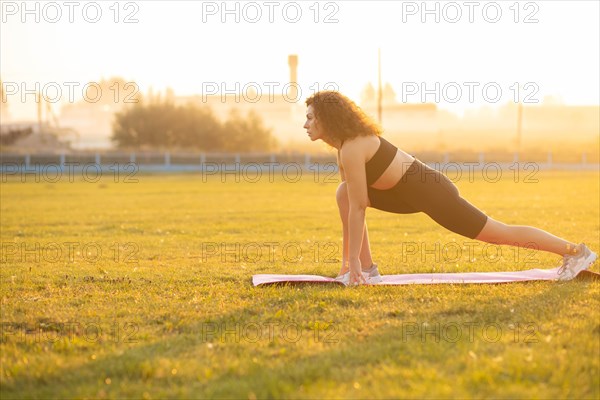 Young curly athletic girl in sportswear doing stretching exercise outdoors on the grass during sunset