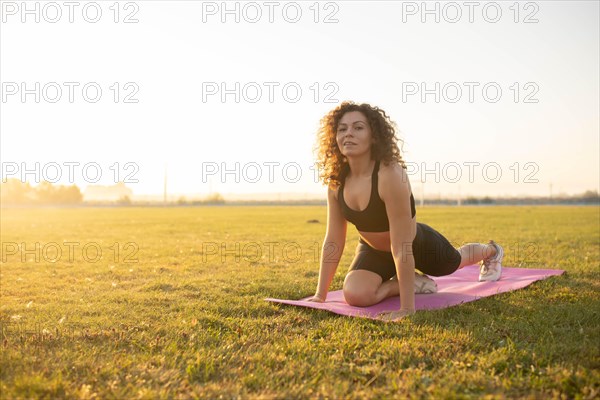 Young curly athletic girl in sportswear doing leg stretching on a yoga mat outdoors on the grass during sunset