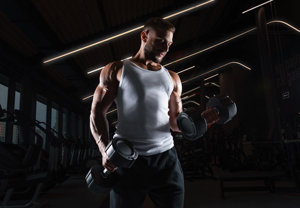 Muscular man in a white t-shirt works out in the gym with dumbbells. Biceps pumping. Fitness and bodybuilding concept.