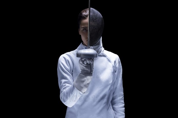 Charming girl in a fencer costume posing on a black background. The sword separates the head into a face and a mask. The concept of fencing.
