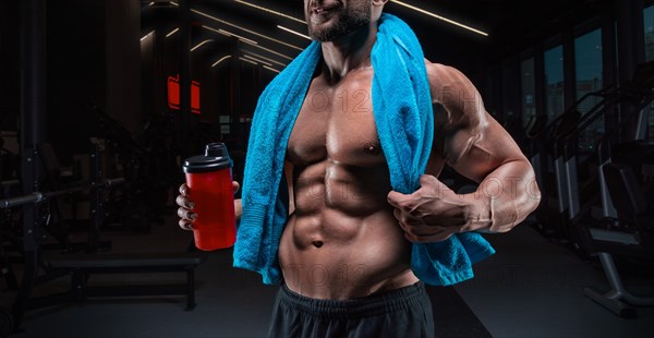 Portrait of a muscular guy with a naked torso and abs. Gym. Blue towel and red shaker. Fitness concept