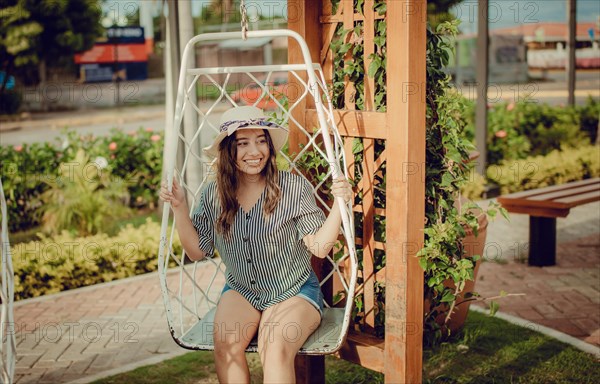 Relaxed girl in hat sitting on a white swing in a beautiful garden. A smiling girl sitting on a swing in La Calzada