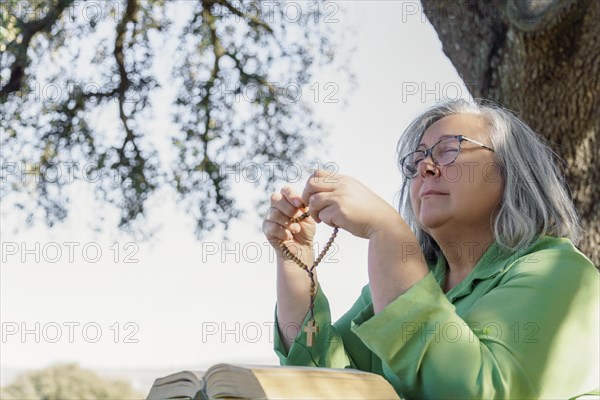 Older white-haired woman with a rosary beads in her hand and a bible praying with her eyes closed sitting at a wooden table in the forest