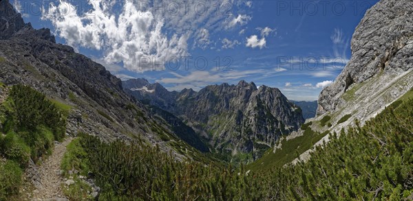 A narrow mountain path winds its way through a landscape of lush greenery and rock faces under a cloudy sky