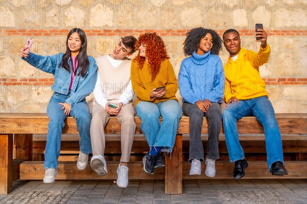 Multi-ethnic young friends taking selfies with the mobile phone sitting on a wood bench in the street