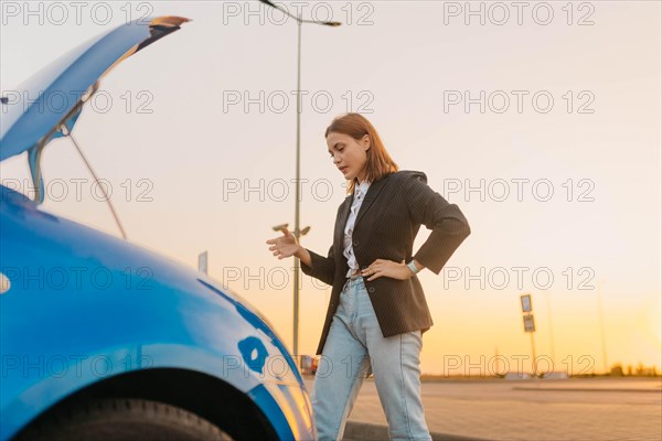 A young beautiful stylish girl driver in a jacket is trying to identify the cause of a car breakdown under the hood