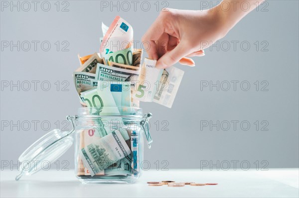 Female hand puts a bill into a glass piggy bank full of money. Thrift concept.