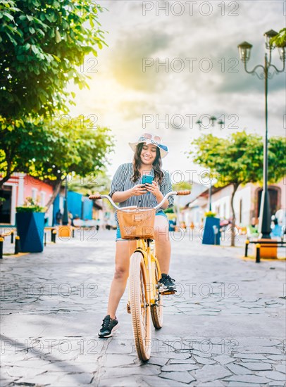 Happy tourist girl on bicycle using cell phone on the street. Beautiful girl in hat on bicycle with cell phone on the street of La Calzada