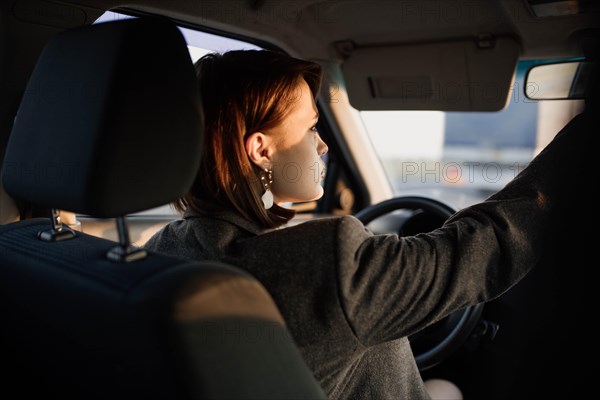 Young beautiful smiling woman taxi driver in a jacket sits behind the wheel of a car and adjusts the distant view mirror