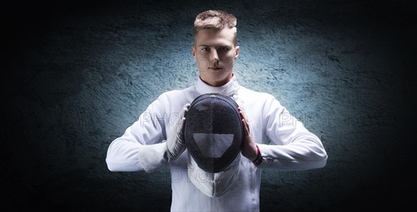 Portrait of a male fencer against a blue spot on the wall. He holds a helmet in front of him. The concept of fencing.