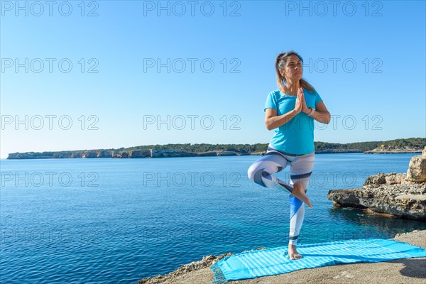 Woman in tree pose practicing yoga on a mat beside a serene sea under a clear blue sky