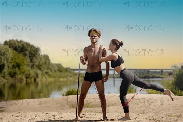 Young athletic couple doing exercise with resistance band on the shore of a lake on a sunny day