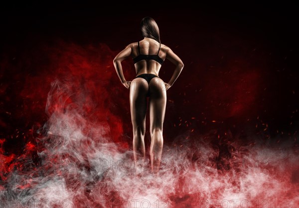 Image of a female athlete in a fire arena surrounded by smoke. Back view. Bodybuilding and fitness concept.