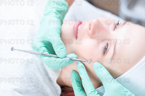 Head of woman lying on couch in white cap in cosmetological clinic with closed eyes and smiling. Doctor holding UNO spoon near her face.