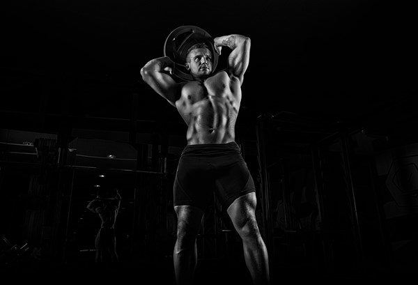 The weightlifter does the workout with burdening. He lifted a heavy pancake over his head and makes circular movements with his hips. Front view