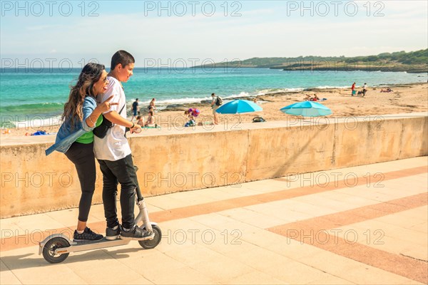 A couple on a scooter enjoying a sunny day at the beach with people and the sea in the distance