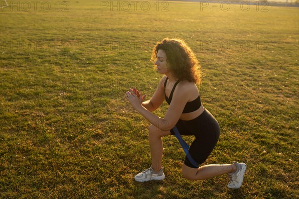 Young curly athletic girl in sportswear performs lunges with resistance band outdoors on the grass during sunset