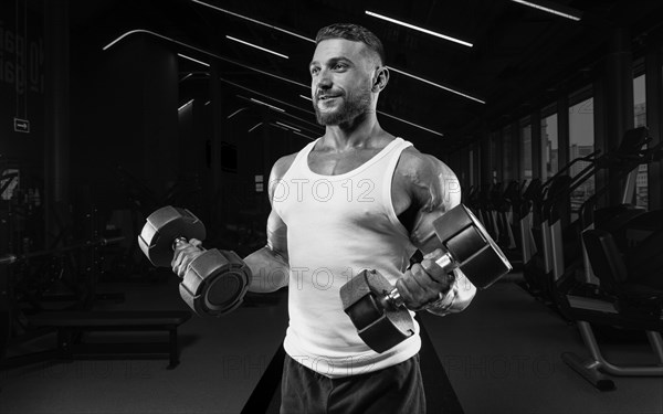 Muscular man in a white t-shirt works out in the gym with dumbbells. Biceps pumping. Fitness and bodybuilding concept.