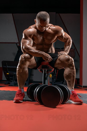 Muscular man sits on a bench near huge dumbbells. Bodybuilding and powerlifting concept.