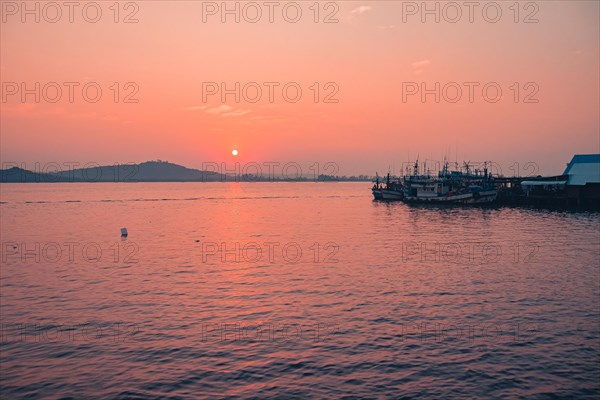 Warm hues fill the sky as the sun sets over a tranquil water scene with silhouetted boats. Koh Sdach Island