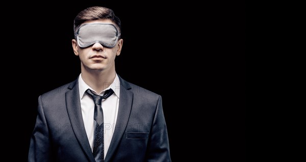 Portrait of a man in a mask for sleeping. He stands against a black wall. Blind business concept. Work during the crisis.