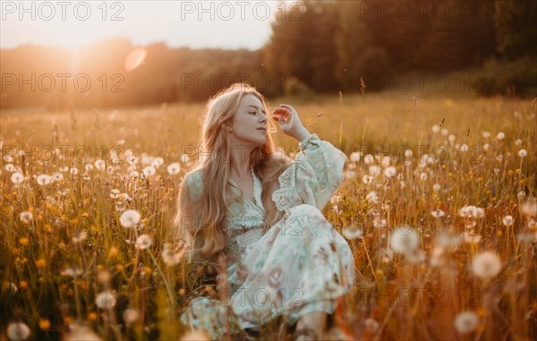 A beautiful young blonde woman with long hair in a white dress walks through a field of dandelions