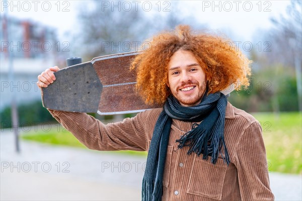 Portrait of a redheaded skater carrying a skate and smiling at camera in an urban park