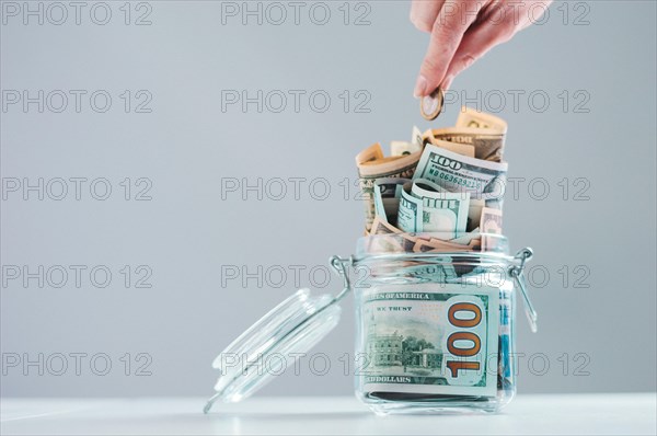 Female hand puts a coin into a glass piggy bank full of money. Thrift concept.