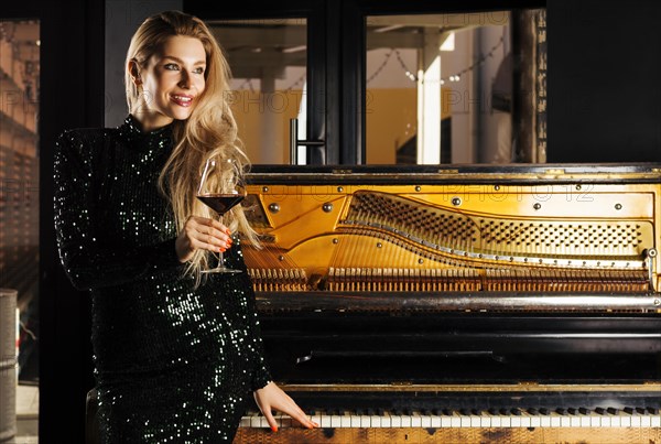 Charming girl in evening dress sits on the piano and poses for the camera with a glass of wine.