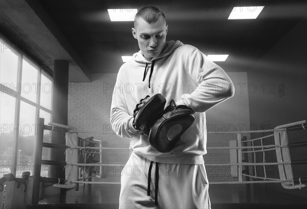 Male trainer in the gym against the background of the ring holds boxing paws. Mixed martial arts concept. High image quality