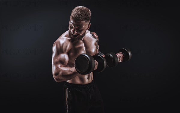 Young muscular guy pumping biceps with dumbbells. Concentrated flexion. Fitness and nutrition concept.