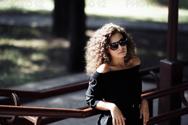 A young slender curly woman in a black dress and sunglasses posing on a sunny day against the backdrop of the city