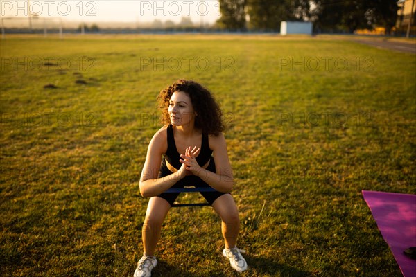 Young curly athletic girl in sportswear performs squats with resistance band outdoors on the grass during sunset