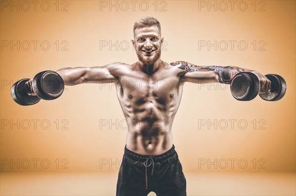 Young muscular guy is pumping deltas with dumbbells on an orange background. Fitness and nutrition concept.