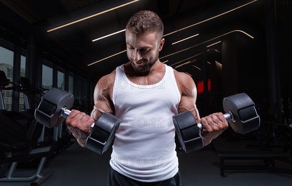 Muscular man in a white t-shirt works out in the gym with dumbbells. Biceps pumping. Fitness and bodybuilding concept.