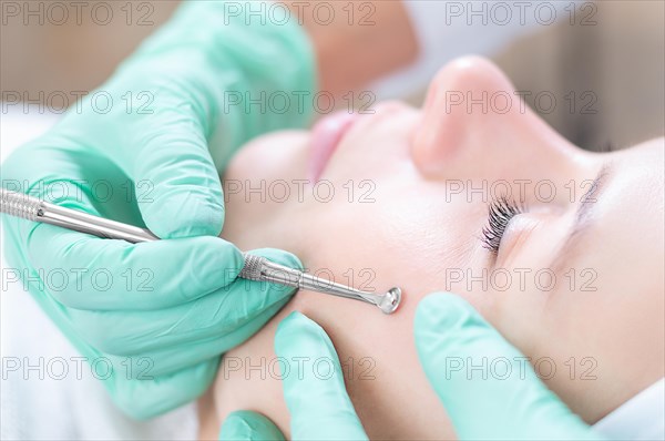 Head of woman lying on couch in white cap in cosmetological clinic with closed eyes and smiling. Doctor holding UNO spoon near her face.