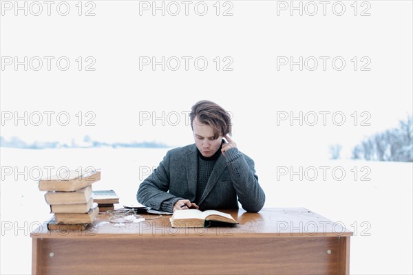 A young handsome intelligent man in a jacket sits at a table in a field and reads ancient books in winter