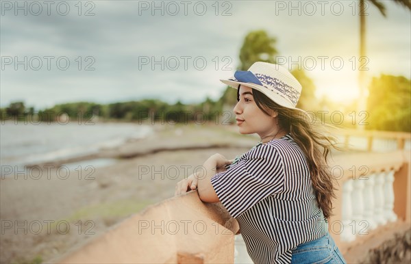 Beautiful tourist girl in hat looking at the beach from a pier. Portrait of female traveler on a pier looking at the horizon
