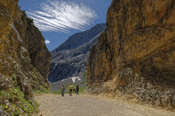A group of hikers on a mountain path surrounded by imposing rocks under a blue sky with foehn clouds
