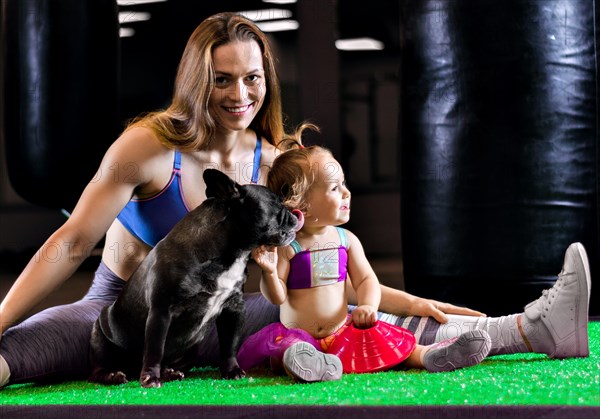 Charming sports mom trains in the gym with her little daughter and a French bulldog.