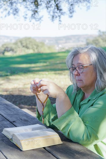 Mature white-haired woman with glasses praying with a rosary beads and a bible on a wooden table in the field