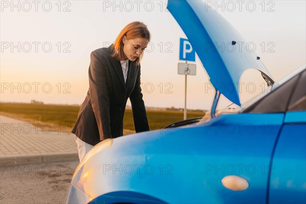 A young beautiful stylish girl driver in a jacket is trying to identify the cause of a car breakdown under the hood