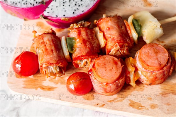 Skewers with bacon-wrapped sausages and vegetables on a wooden cutting board with dragon fruit